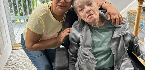 A middle aged adult with short hair and glasses embraces a senior adult using a wheelchair. They are posing for a picture in a home's hallway.