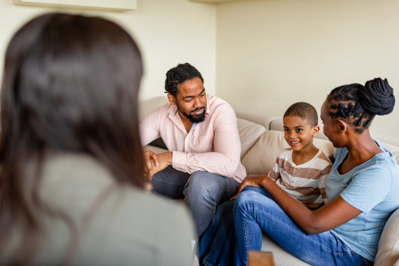 Two parents and a young child sit on a couch smiling at a therapist