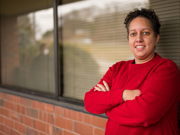 Smiling person in red shirt standing with arms crossed in front of a brick wall