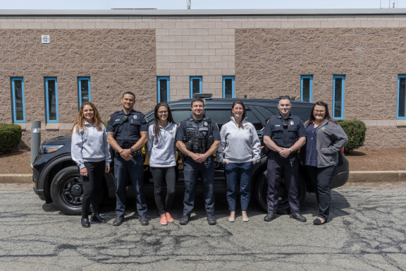 Seven adults standing in front of a police SUV cruiser, smiling at the camera. Three are in law enforcement uniforms.