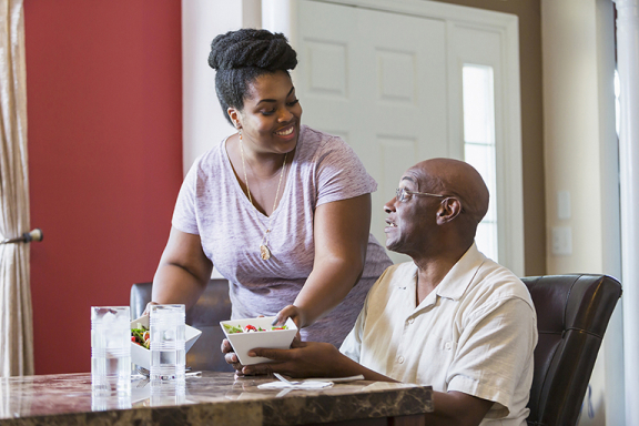 Young adult woman serving a meal to an older man at the kitchen table.