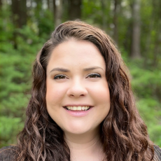 An adult with long curly brown hair smiles at the camera