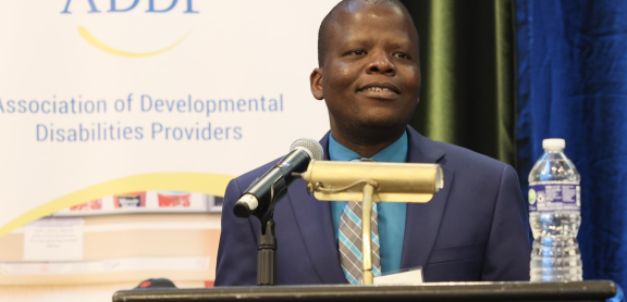 An adult with brown skin and a shaved head wears a suit behind a podium. He is smiling.