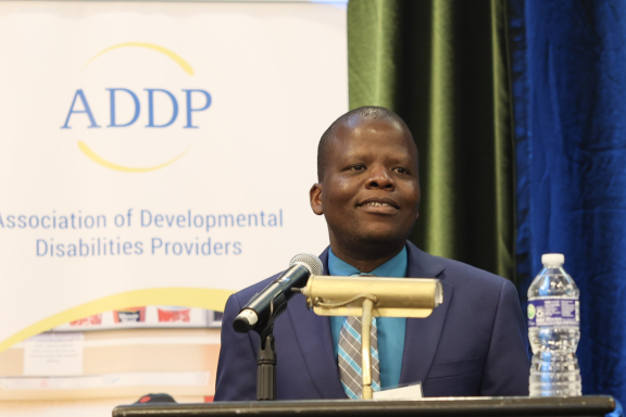 An adult with brown skin and a shaved head wears a suit behind a podium. He is smiling.