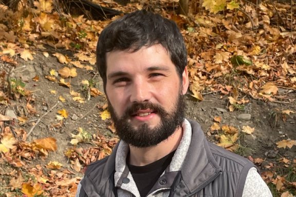 A man with light skin and dark hair smiles at the camera in a fall outdoor setting.